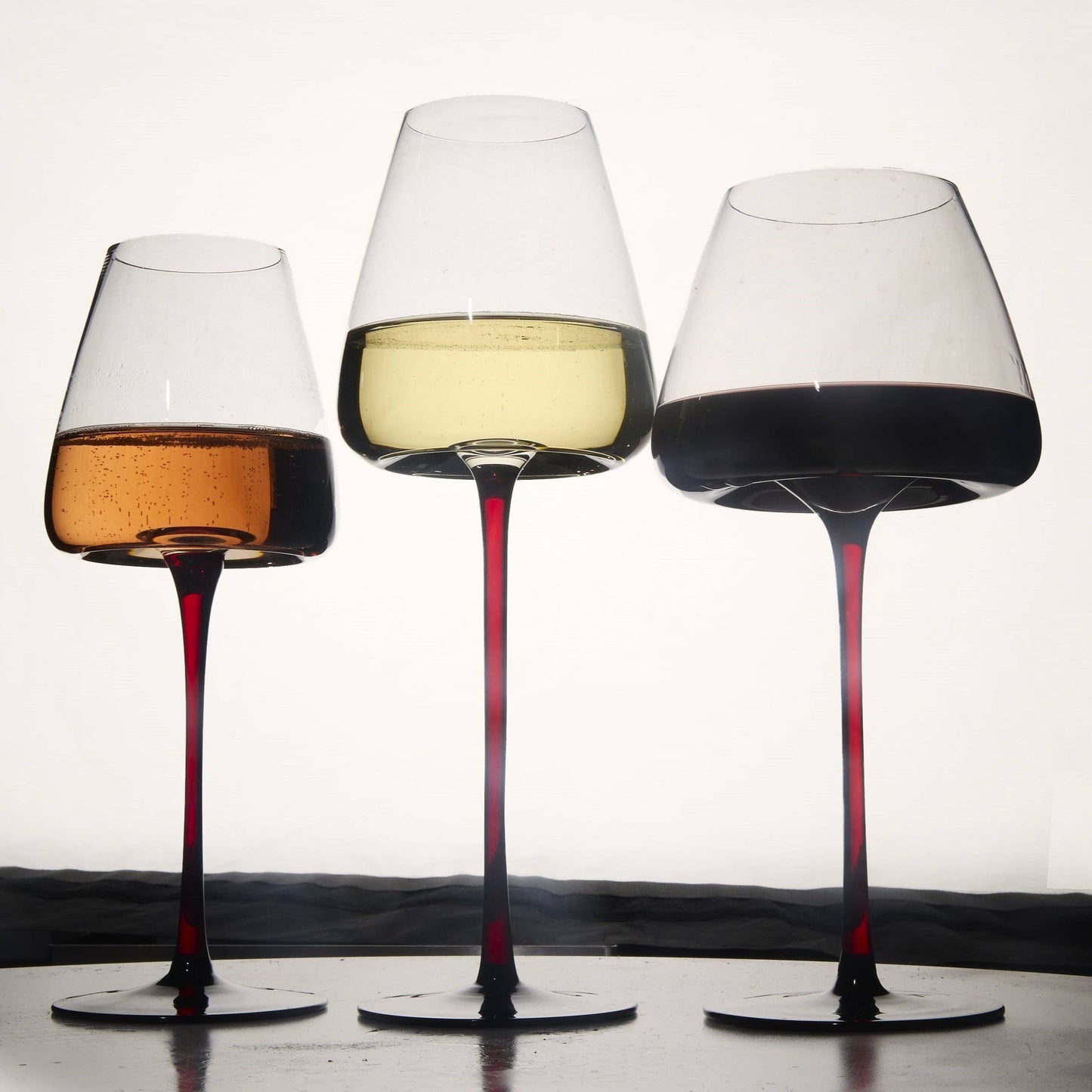 Three 24oz extra-large concave wine glasses with red stems, holding amber, pale yellow, and deep red wine.