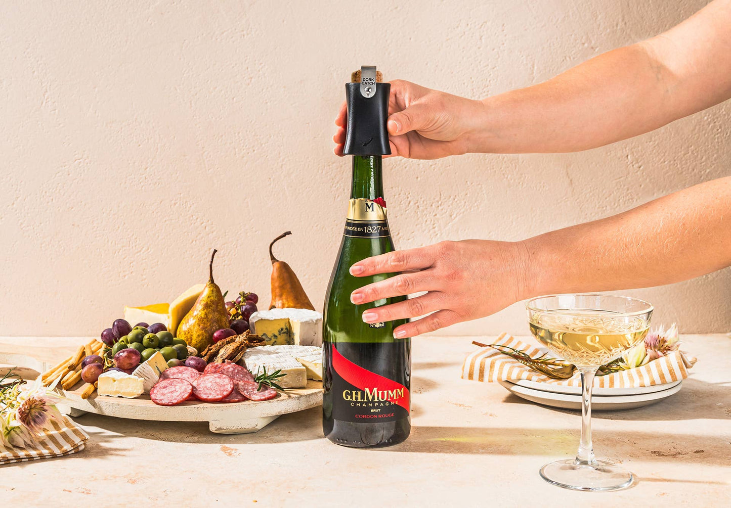 Hands operate Champop champagne popper to safely pop and catch the cork beside a cheese board and champagne flute.