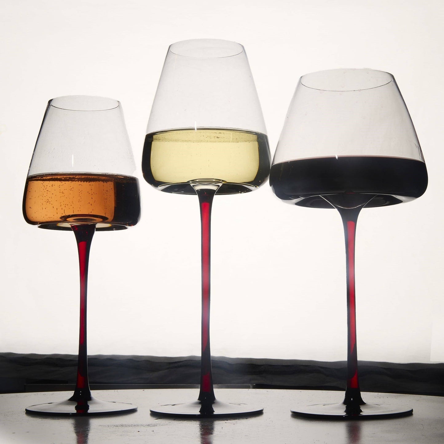 Three 16oz concave wine glasses with ombre red stems, each holding wine of different colors.
