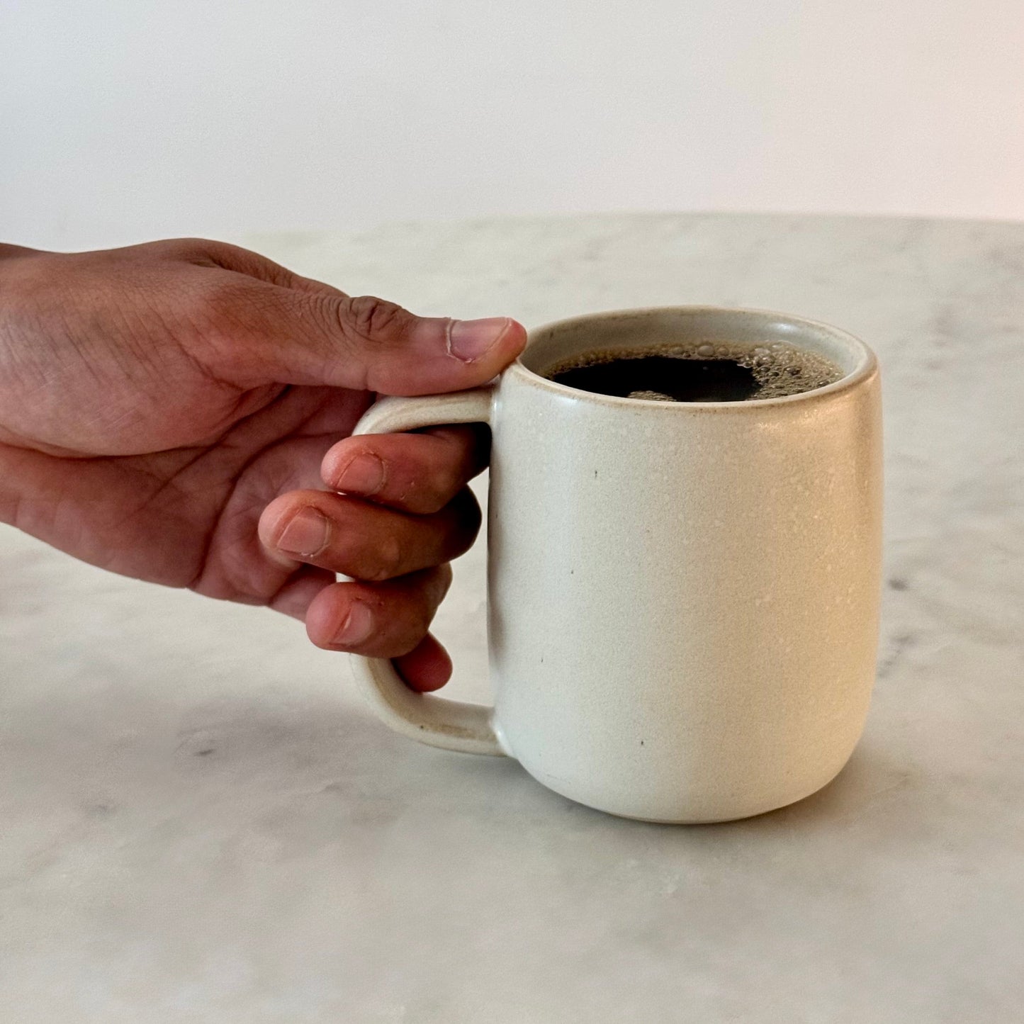 A hand holds The Standard Mug, a 12oz off-white ceramic mug filled with coffee on a marble countertop.