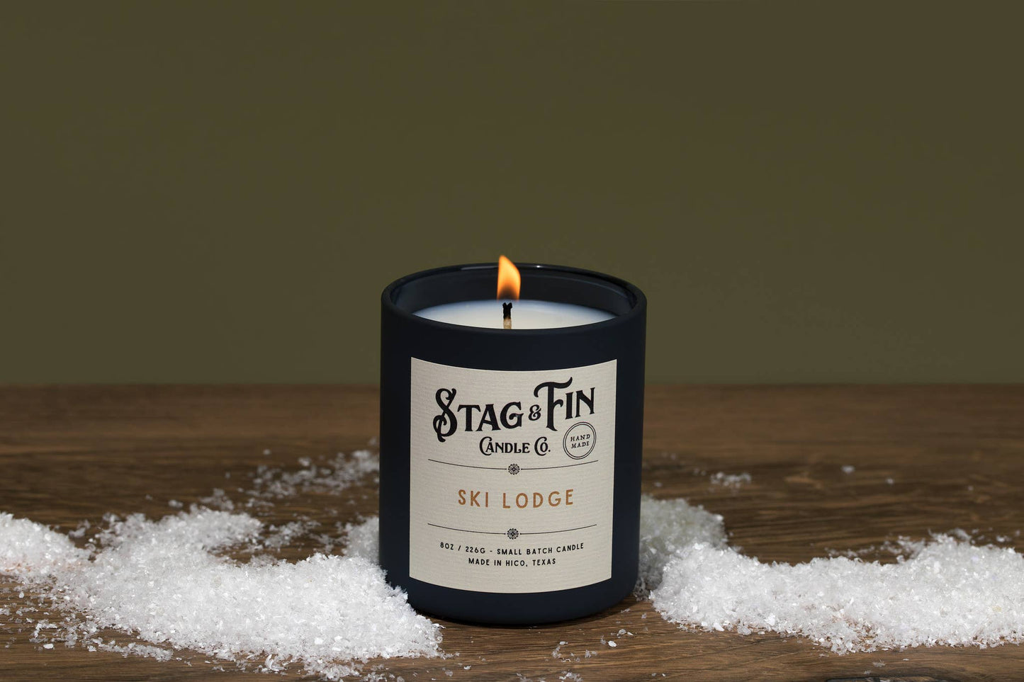 Ski Lodge candle by Stag & Tin Candle Co. in a dark jar with cream label, lit wick, snow on wood.