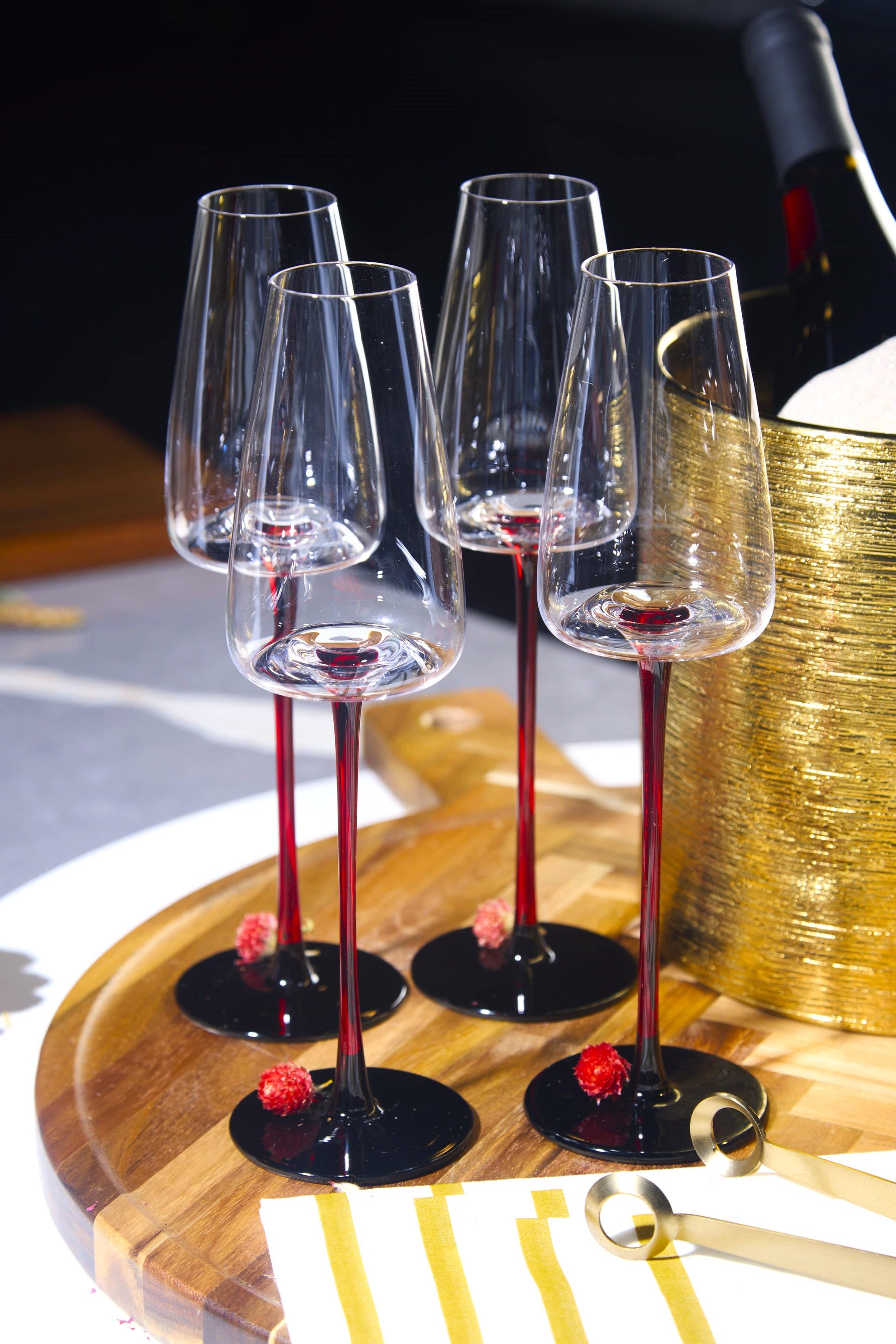Four concave 9oz champagne glasses with red stems on a wooden tray beside a gold champagne bucket.