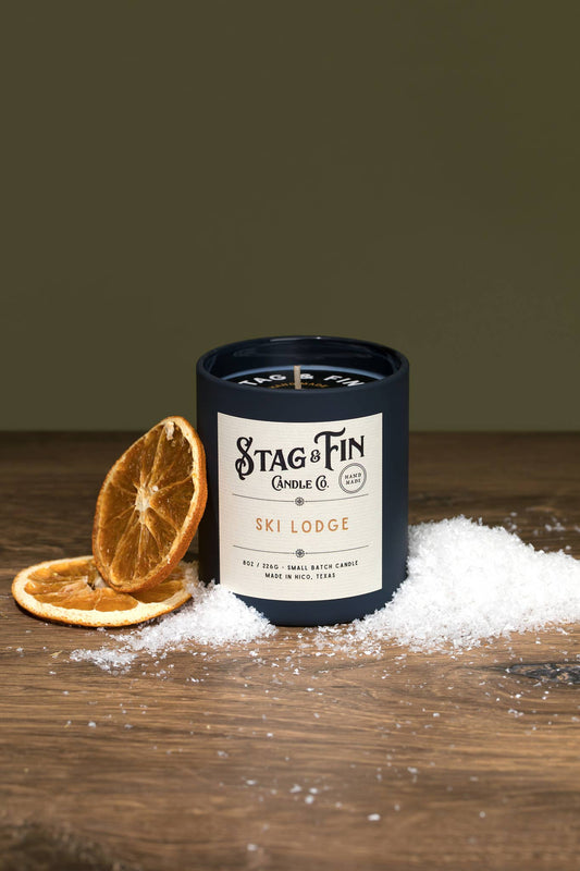 Ski Lodge candle in a navy jar with dried orange slices and snow on a wooden table.
