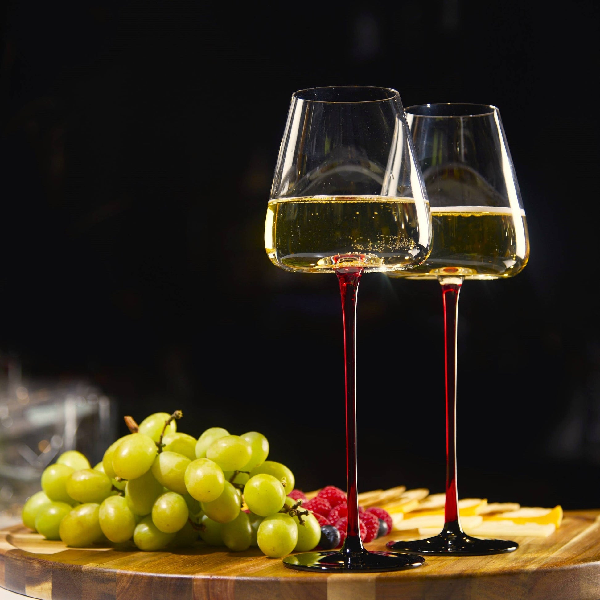 Two 16oz concave wine glasses with ombre red stems on a wooden board beside grapes and cheese.
