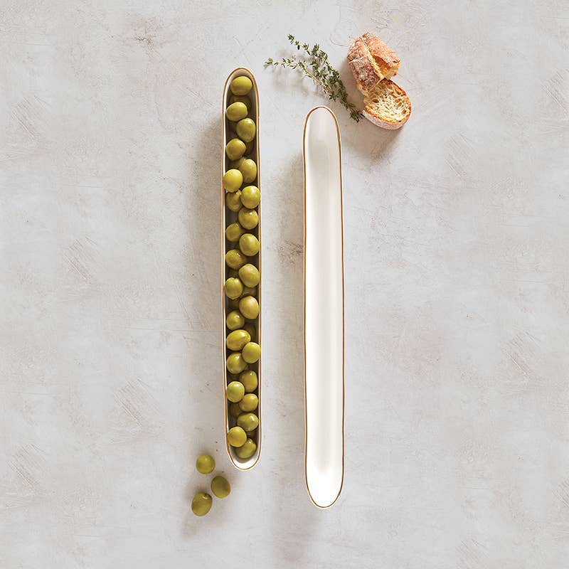 Two slim white ceramic olive boats, one filled with green olives, with bread slices and thyme nearby.