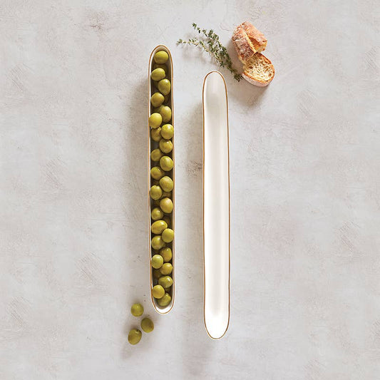 Two slim white ceramic olive boats, one filled with green olives, with bread slices and thyme nearby.