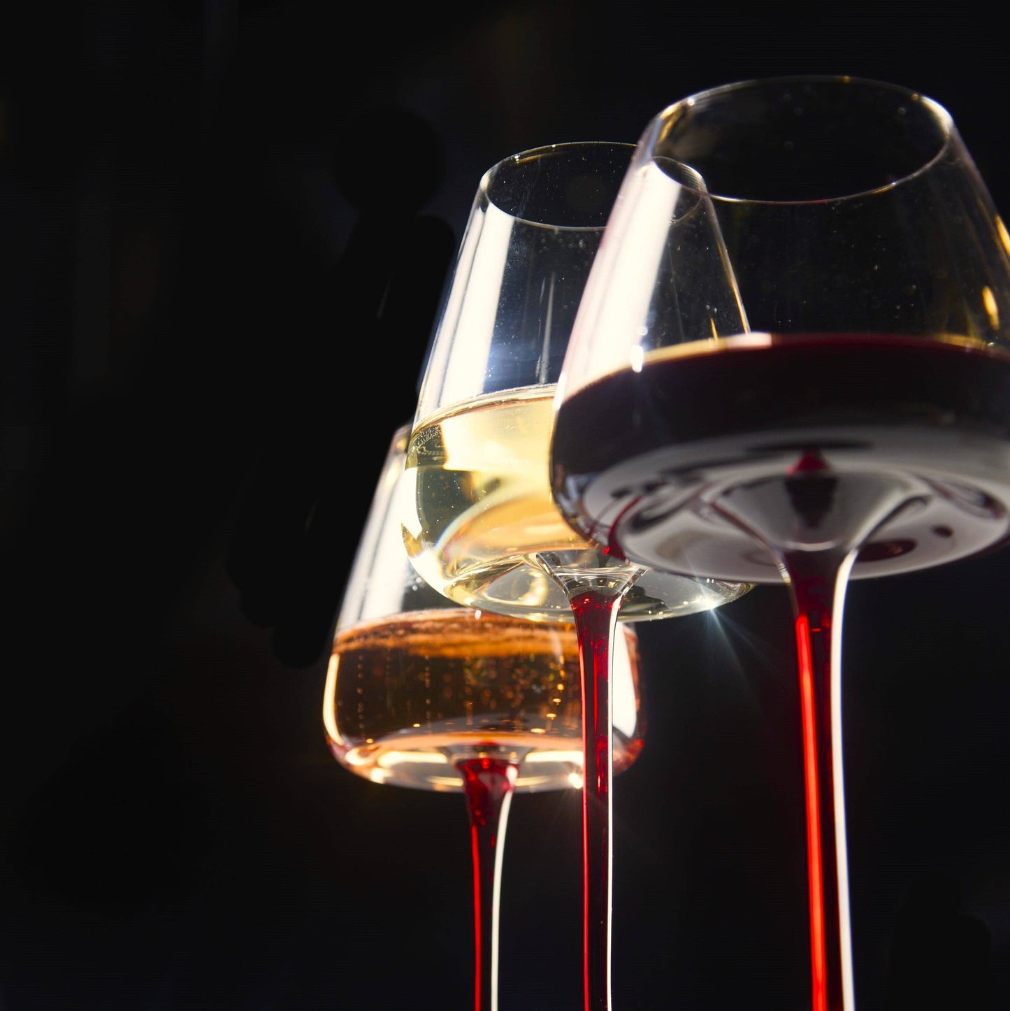 Two concave wine glasses with ombre red stems, holding white and red wine, set against a dark background.