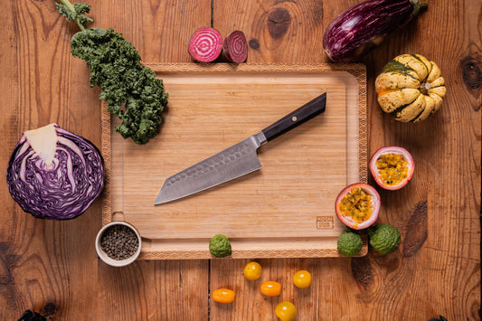 KOTAI Bunka Santoku kitchen knife on cutting board with kale, red onion, cabbage, passion fruit, and tomatoes.