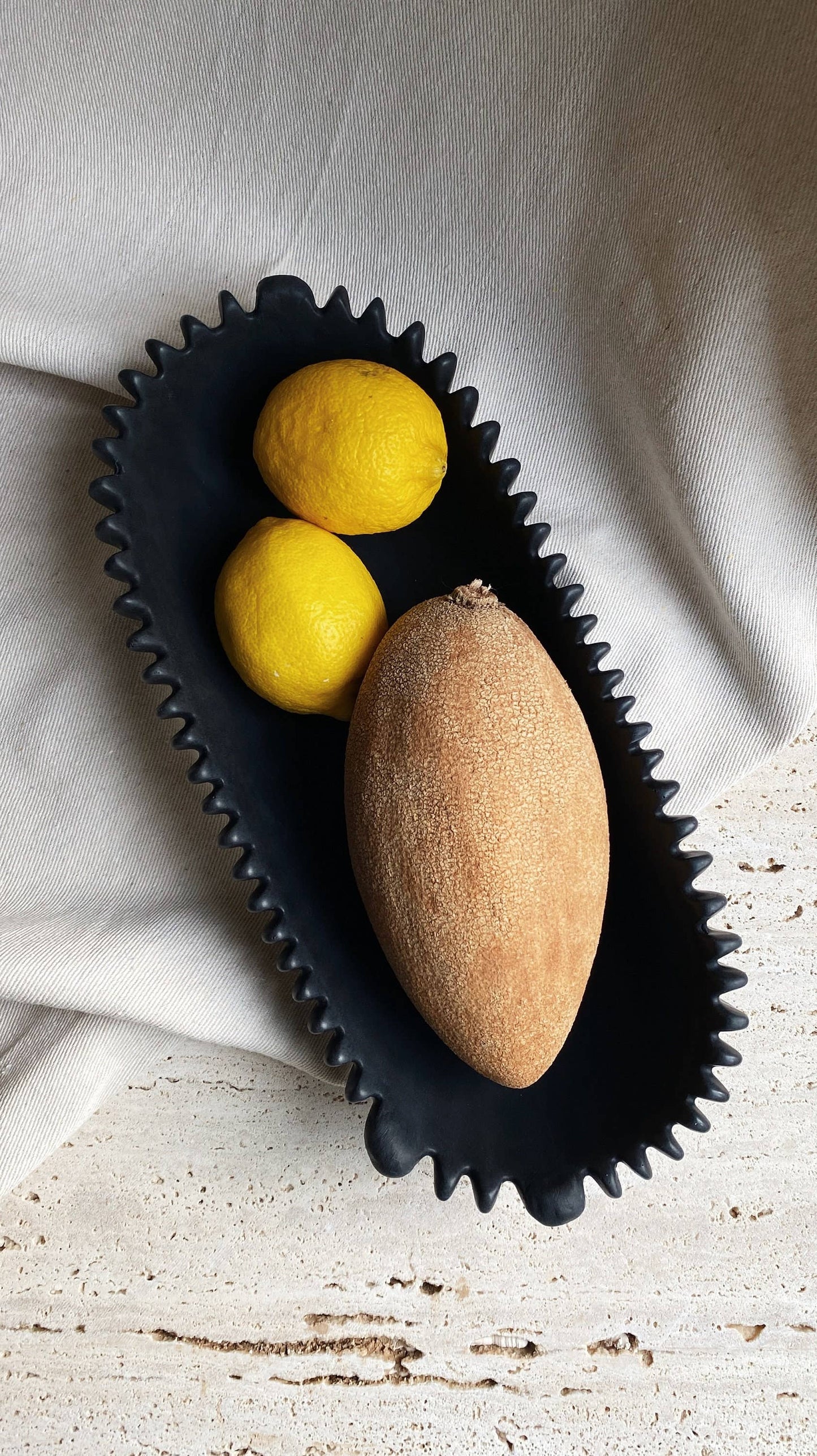 FORMA handmade black oval concrete bowl with a scalloped edge, holding two lemons and a large brown fruit.