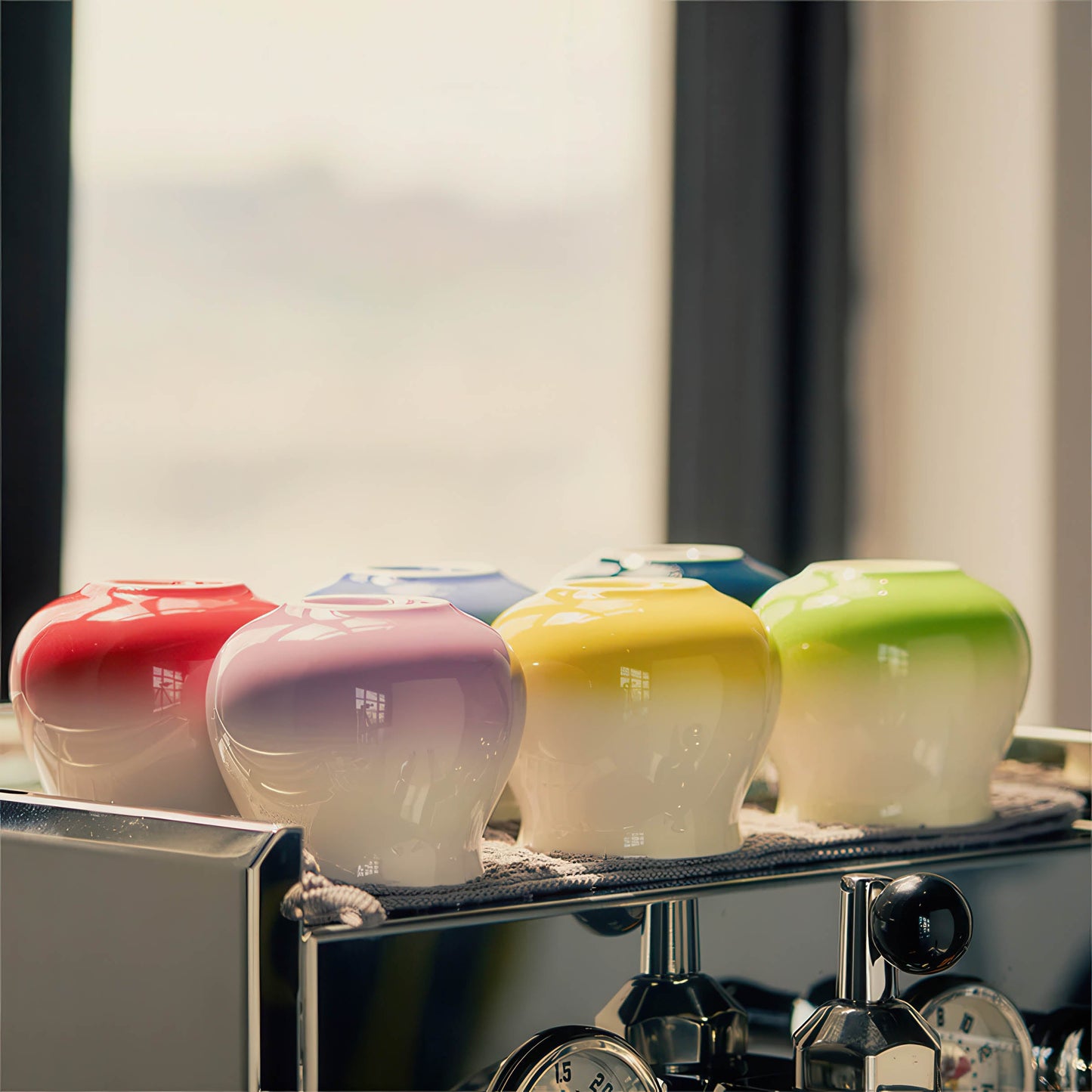 Set of gradient ceramic coffee cups in red, purple, yellow, and green atop an espresso machine