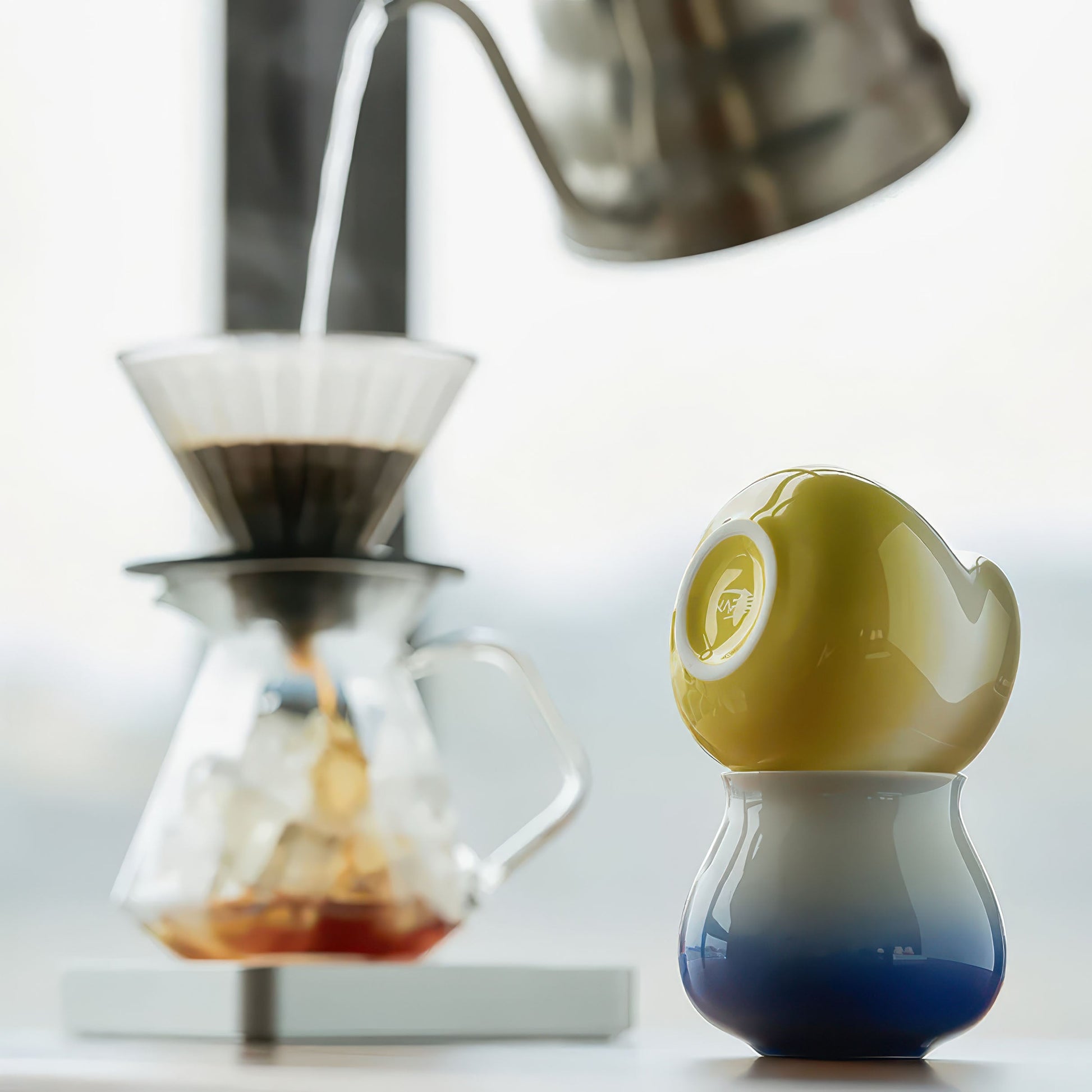 Ceramic gradient coffee cup with blue base and white body, yellow lid, beside a pour-over setup.
