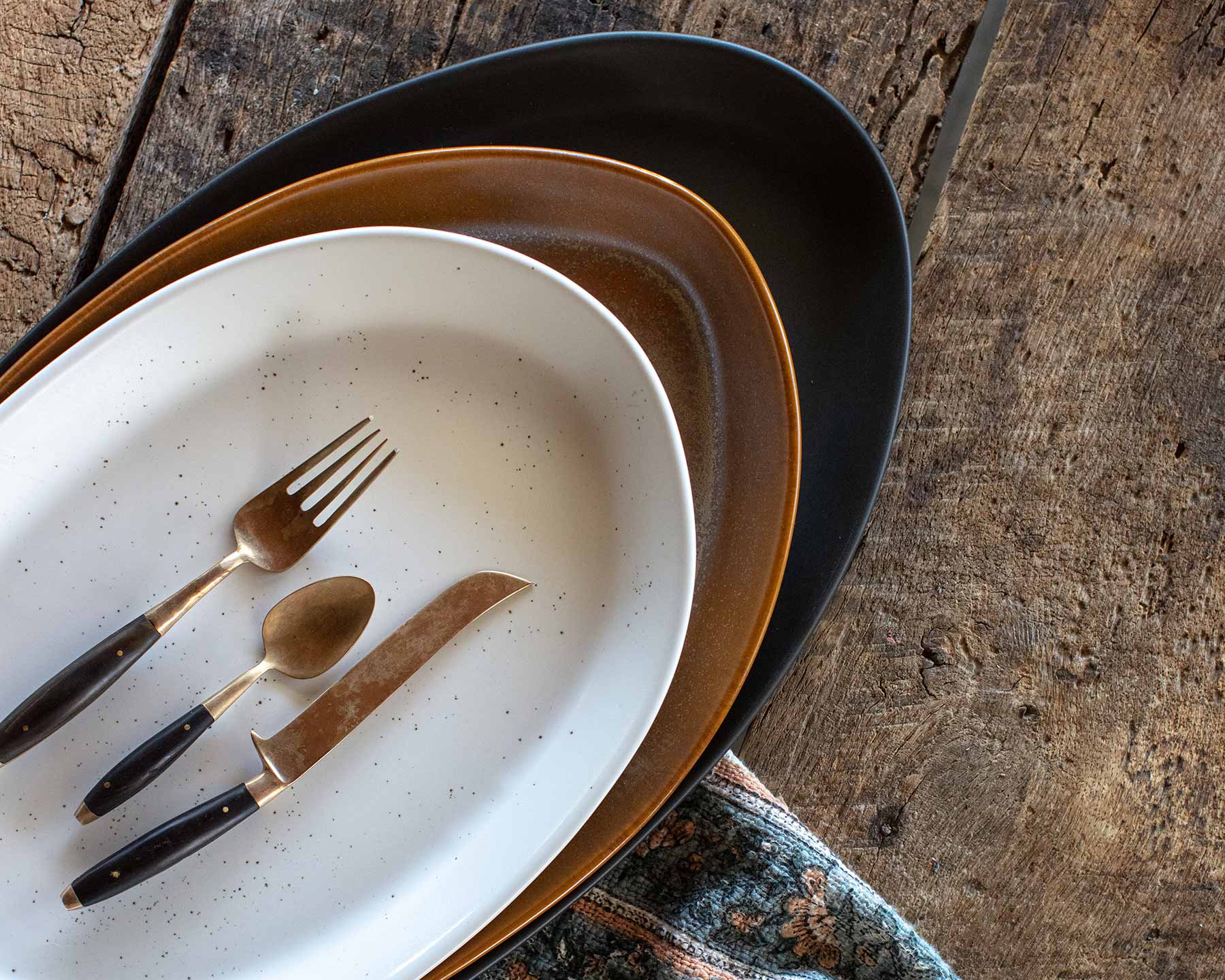 Three oval serving platters set in white speckled, brown, and black, with copper-handled cutlery on a wood table.