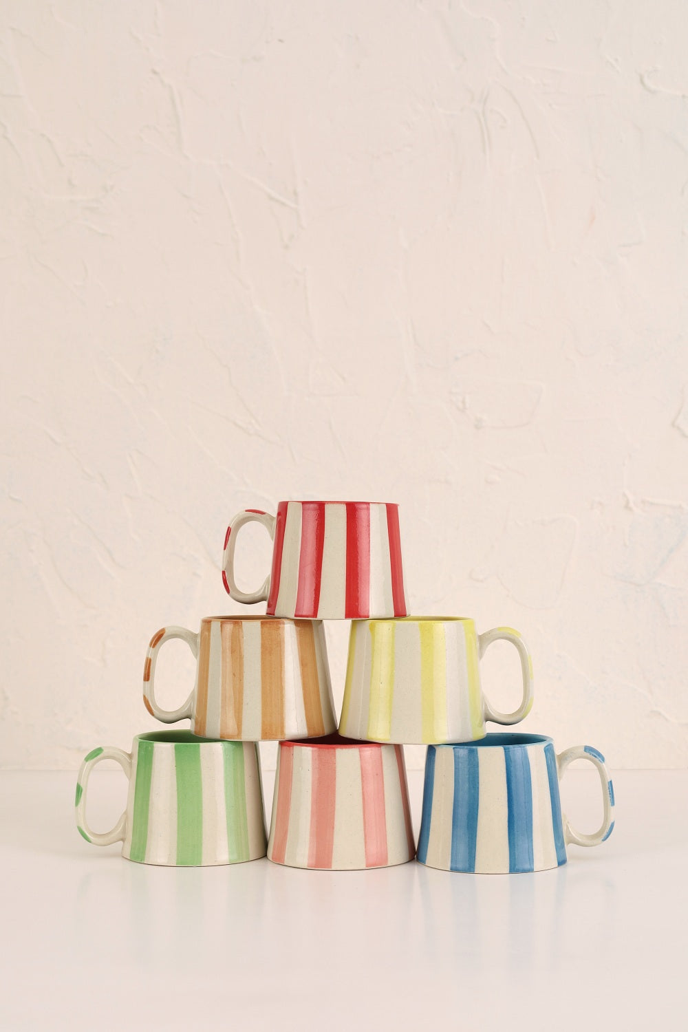 Four ceramic coffee cups with pink stripe pattern, stacked in pastel hues on a white surface.