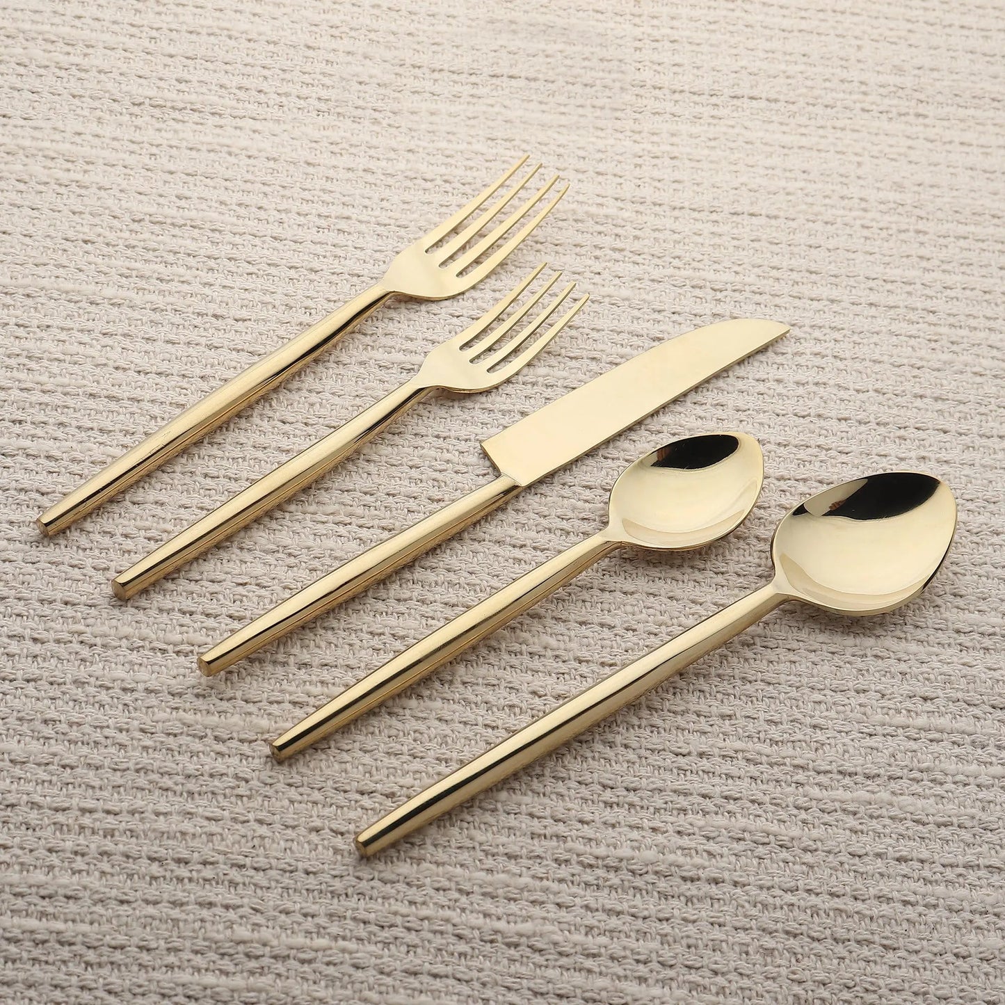 Brio PVD gold 5-piece flatware set with slim tapered handles on beige fabric.