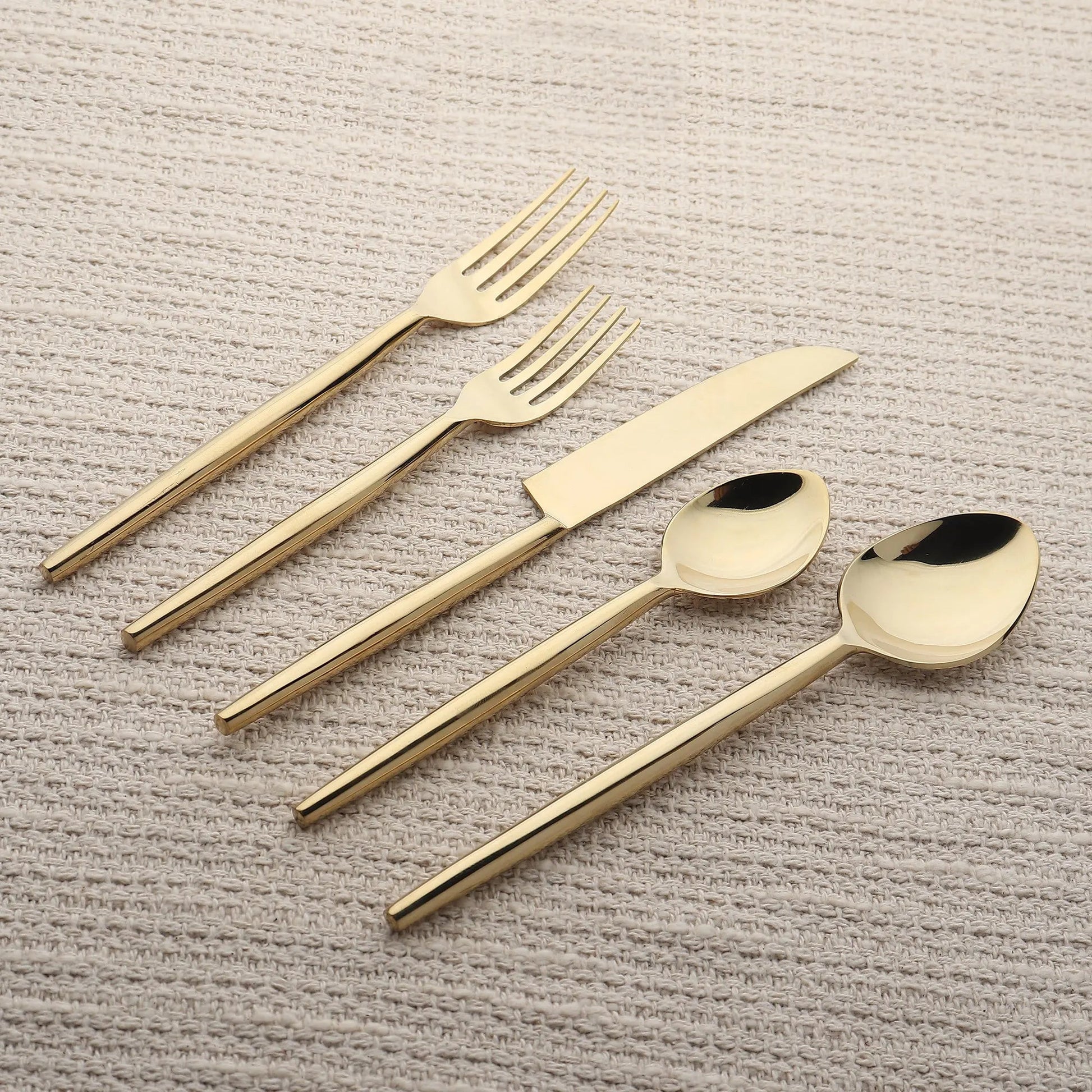 Brio PVD gold 5-piece flatware set with slim tapered handles on beige fabric.