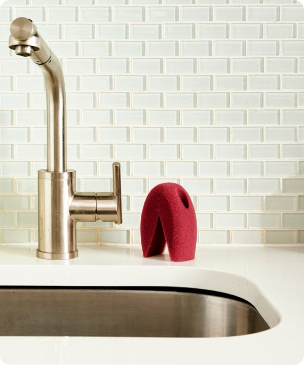 The Three-Up sponge beside a stainless steel kitchen faucet on a white countertop.