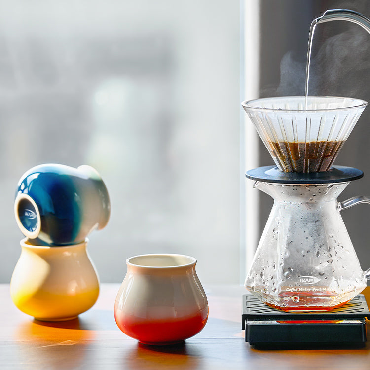 Ceramic Gradient Coffee Cup with orange-red glaze beside a pour-over coffee setup.