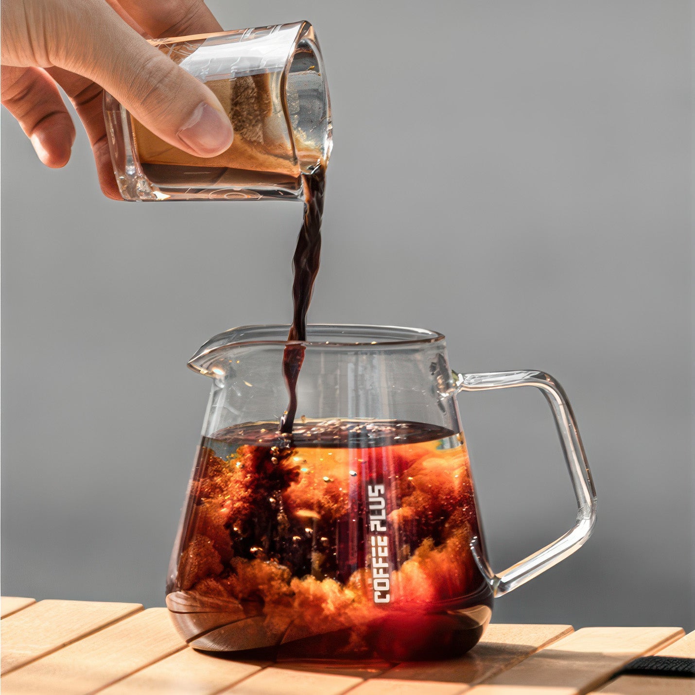 Hand pours hot coffee into a clear glass coffee server carafe for pour-over on a wooden table.