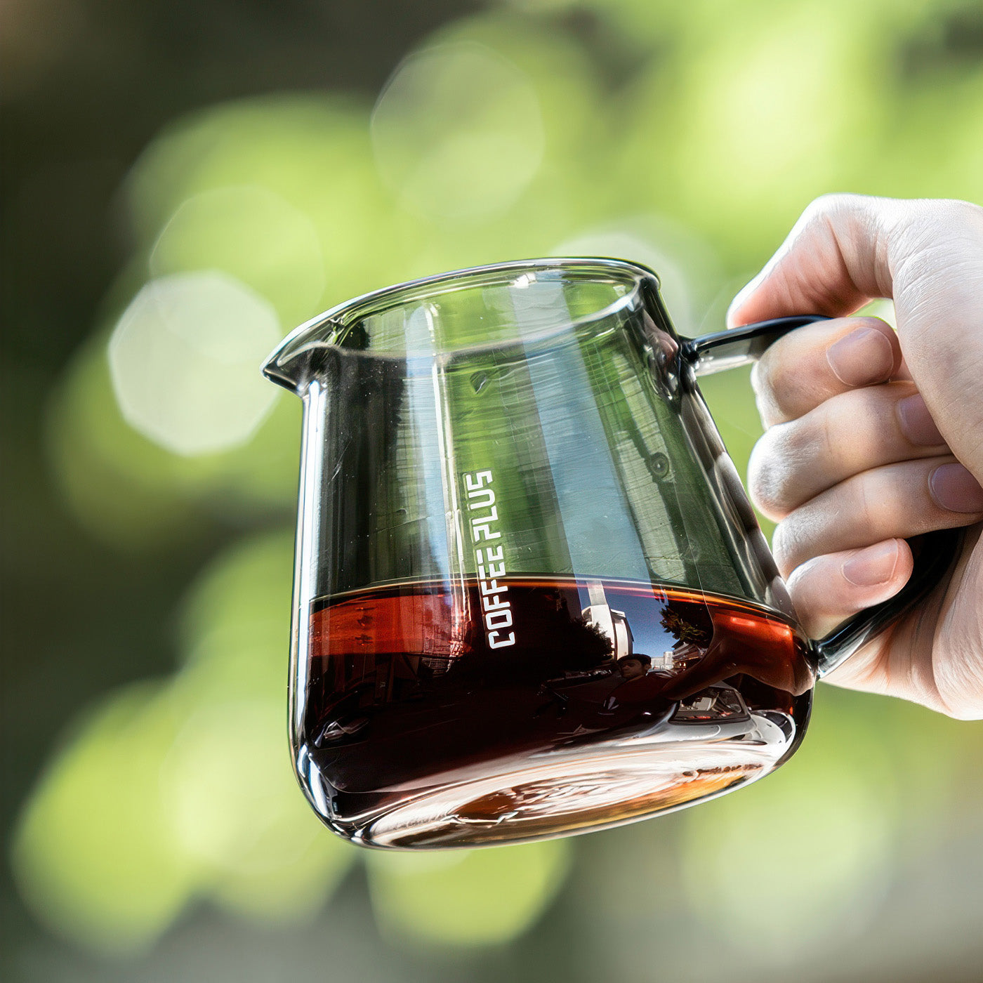 Glass coffee server carafe held in hand for pour-over brewing, clear borosilicate glass with coffee, outdoor green bokeh.
