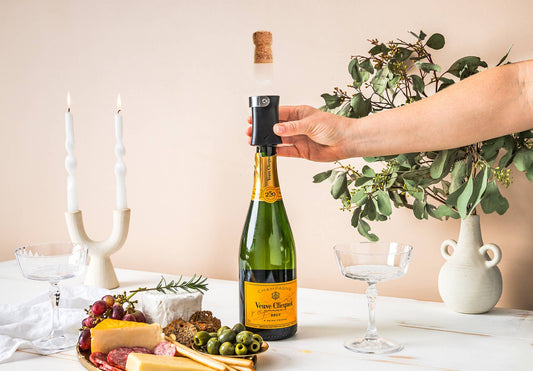 Champop - Champagne Popper & Cork Catcher in use, catching the cork as a hand opens a bottle, with cheese board nearby.