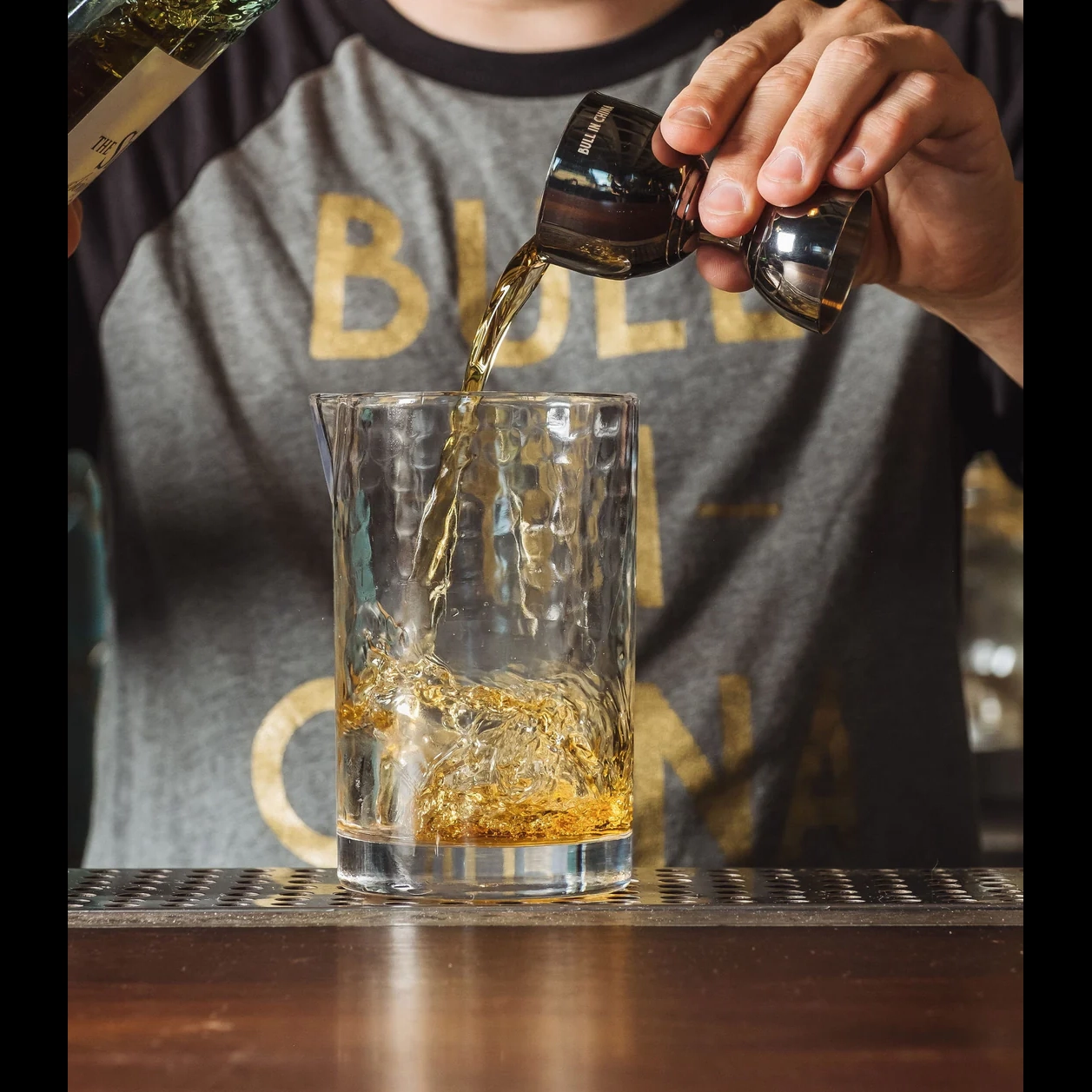 Pouring golden whiskey into BULL IN CHINA The Flagship Mixing Glass with hammered texture.