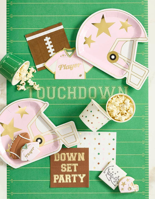 Green football field table runner with yard lines and TOUCHDOWN text, styled with pink helmet plates, football napkins, and party snacks.
