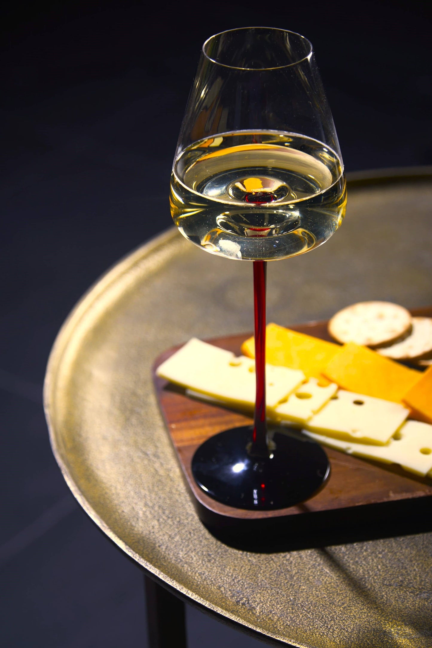 Large concave wine glass with ombre red stem, on a cheese board tray with white wine.