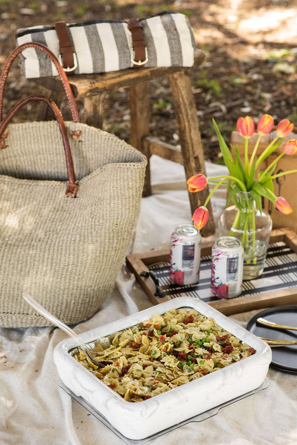 Marble pan frame holding foil pasta salad with serving spoon at outdoor picnic, next to woven bag and tulips.