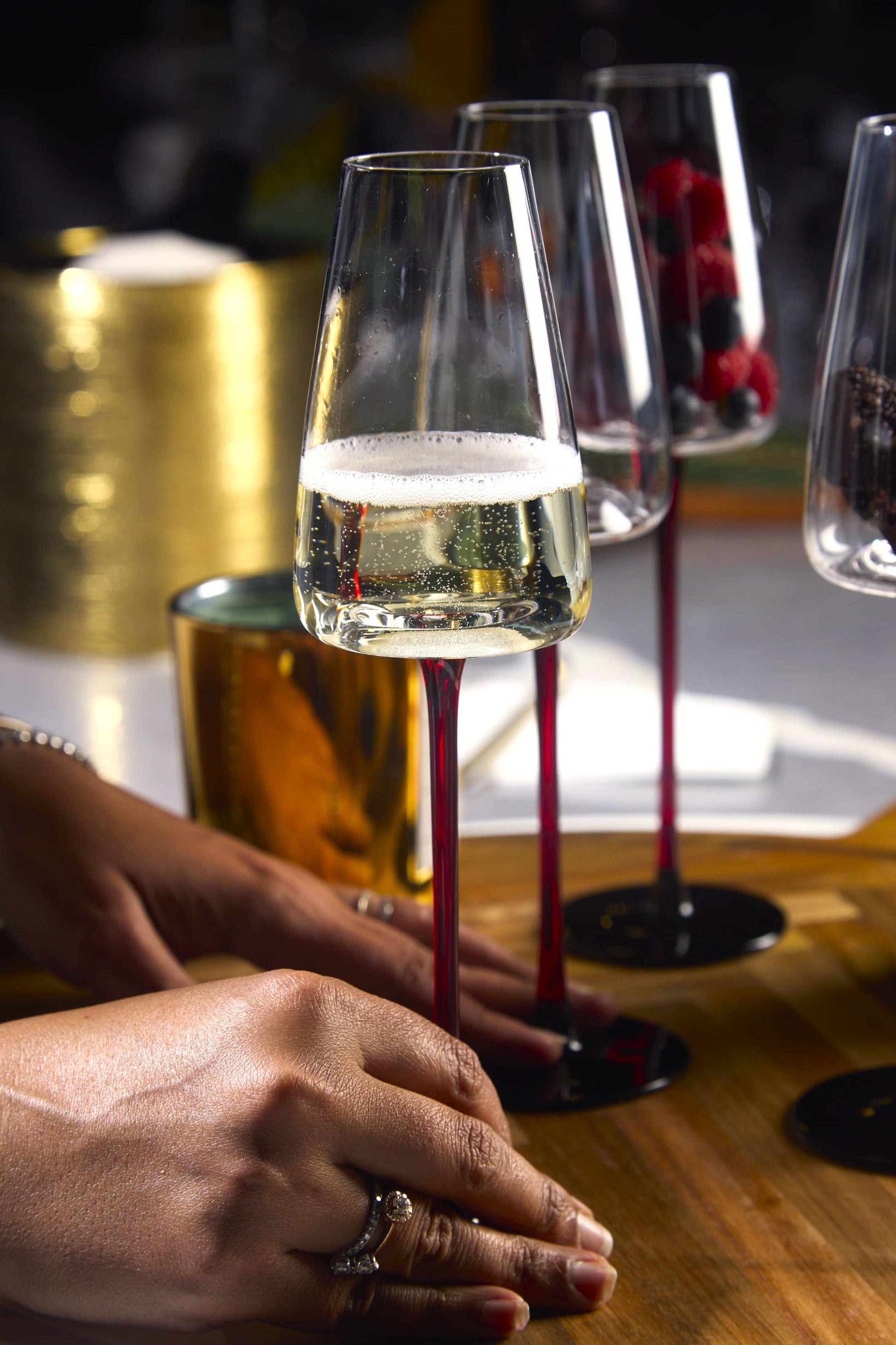 Concave champagne glass, 9oz, with red stem, held by hands on a wooden table; other glasses in background.