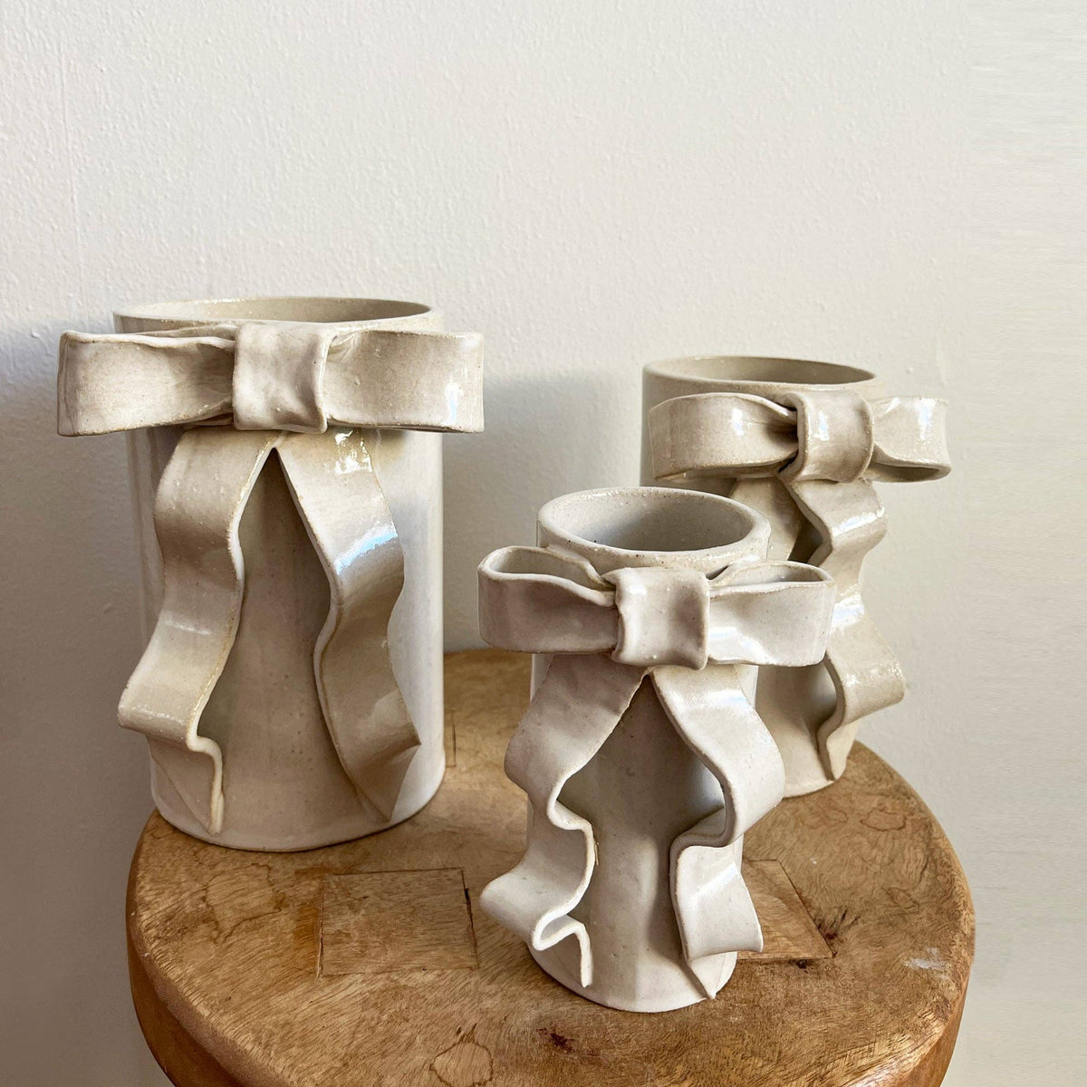 Handmade ceramic vases featuring sculpted ribbon bows in glazed white stoneware. Cylindrical design adds whimsical charm to floral arrangements.