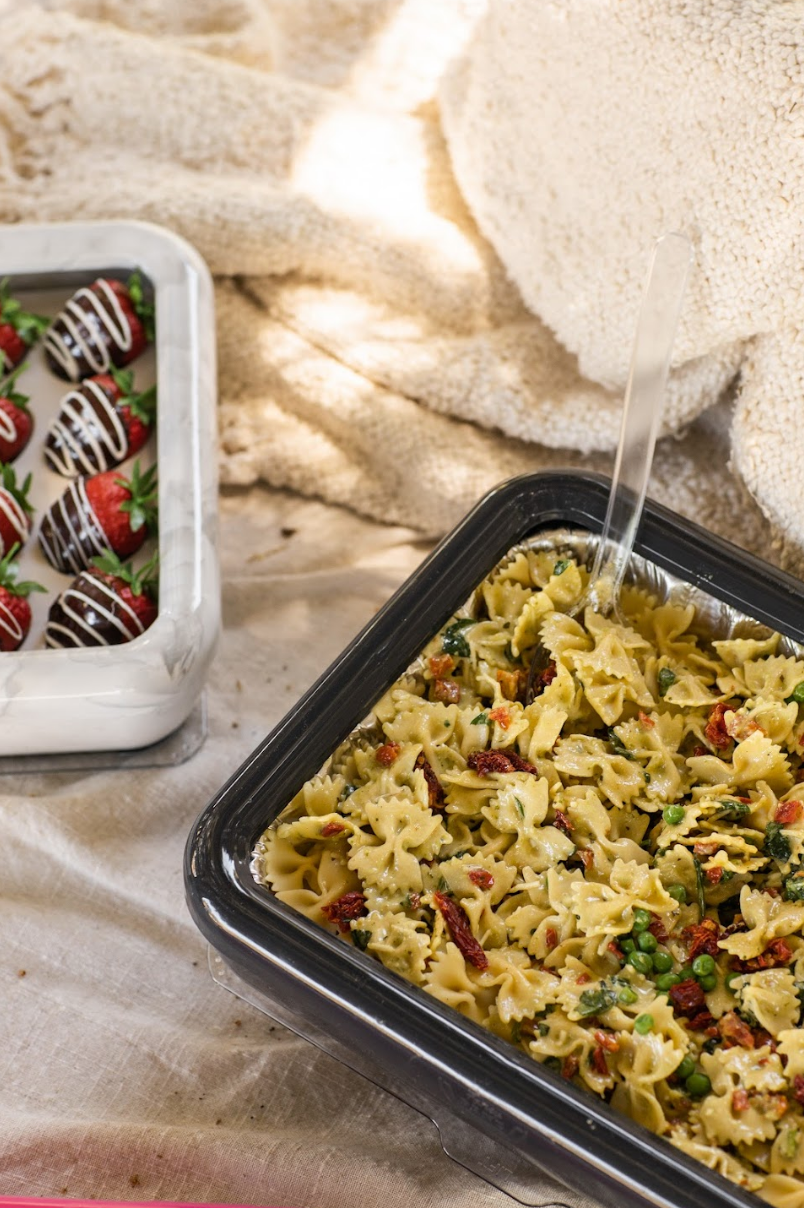 Marble pan frame carrier holding bowtie pasta salad with clear spoon, alongside chocolate-covered strawberries in a matching tray.