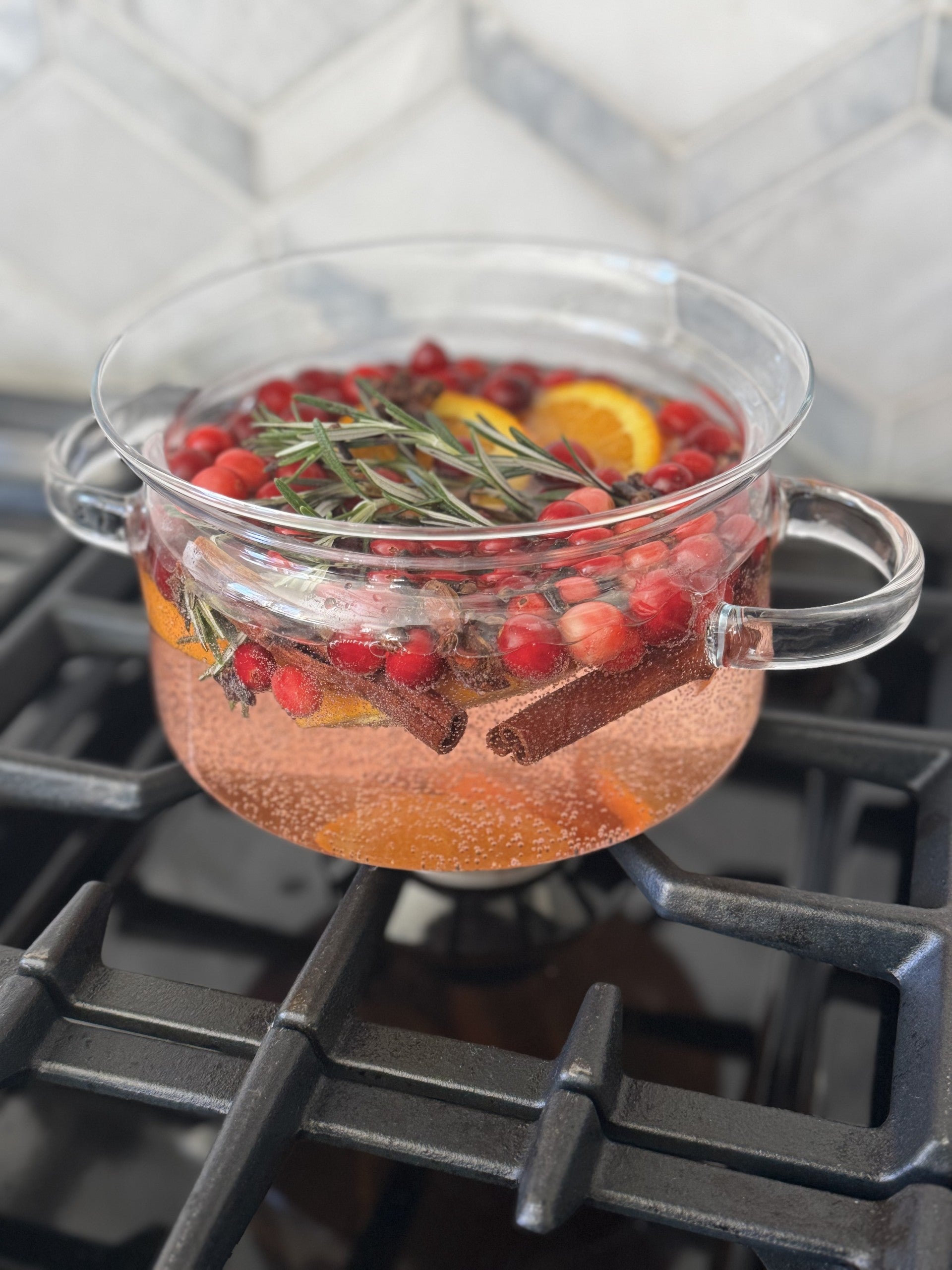 1.5L clear glass simmer pot with lid on stove, filled with cranberries, orange slices, rosemary, and cinnamon.